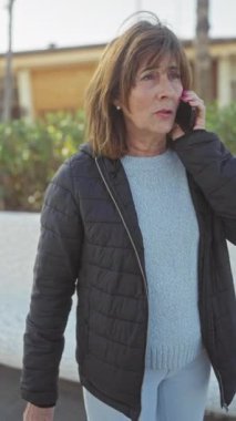 Açık havada akıllı telefondan konuşan, ceketli ve kazaklı olgun bir kadın.