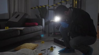 Sakallı orta yaşlı bir adam, olay yerinde kanıt fotoğrafı çekiyor, eldiven ve ayakkabı kılıfı giyiyor..