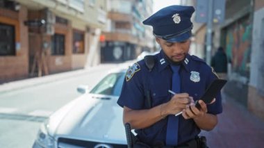 Odaklanmış bir Afro-Amerikan polisi şehir arka planında devriye arabasıyla bir not defterine yazı yazıyor.
