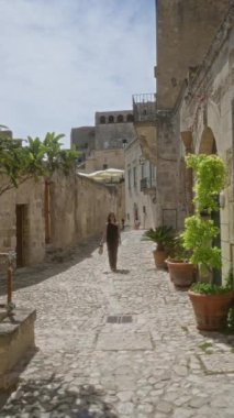 Genç bir kadın, kırsal taştan binalar ve bereketli saksı bitkileriyle çevrili, Materya, Basilicata, İtalya 'daki eski bir kasabanın kaldırımlı sokaklarında yürüyor. Açık havada güneşli bir günün tadını çıkarıyor..