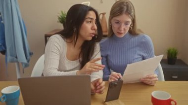 Masada dizüstü bilgisayarı ve akıllı telefonu olan iki kadın oturma odasında belgeleri inceliyor..