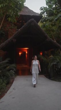 Beyaz elbiseli gülümseyen bir kadın, alacakaranlıkta yemyeşil yapraklarla çevrili egzotik bir bali otelinin önünde duruyor..