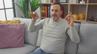 Orta yaşlı sakallı bir adam, rahat bir kazak giyiyor, evde yoga yapıyor, gözleri nazikçe kapanıyor, sakin bir meditasyon hareketi sergiliyor..