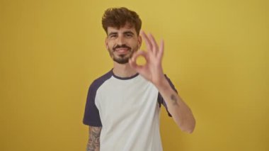 Neşeli, genç İspanyol bir adam izole edilmiş sarı arka planda duruyor, mutlu bir el hareketi yapıyor, büyüleyici bir gülümsemeyle parmaklarının arasından bakıyor.