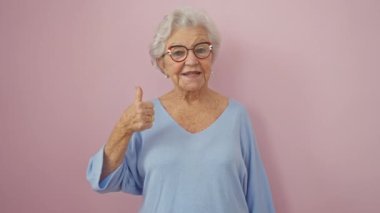 Neşeli, gri saçlı, son sınıf öğrencisi kadın kendine güvenerek tamam işareti gösteriyor, neşe ve onayı ifade ediyor, pembe arka planda duruyor, pozitif, başarılı bir yaşam tarzıyla ışıldıyor..