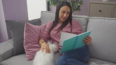 İspanyol kadın kanepede kitap okuyor. Sıcak bir oturma odasında Malta köpeğiyle.