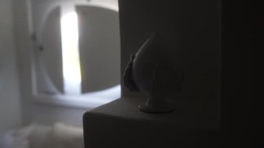 Beyaz süslemeli bir heykel, arka planda kısmen açık bir pencere perdesinden süzülen ışığın olduğu bir çıkıntının üzerinde oturuyor.