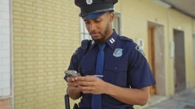 Şehir sokağında dikilirken akıllı telefon kullanan üniformalı siyahi bir polis..