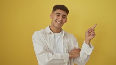 Tişörtlü neşeli genç Arap adam, tutkuyla parmaklarıyla kenarı işaret ediyor, gülümsüyor, canlı sarı bir arkaplan karşısında duruyor.!