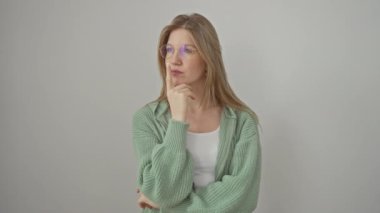 Gözlük takan konsantre genç sarışın kadın, merak ve şüphe içinde duruyor, parmağı çenesinde, izole edilmiş beyaz bir arka plana dikkatlice bakıyor.