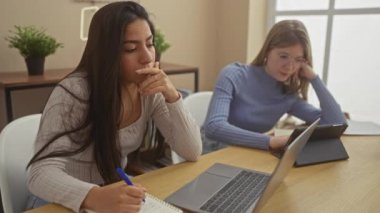 İki kadın modern bir ev ofisinde odaklanmış ve birden fazla iş yapıyor, biri dizüstü bilgisayarda çalışıyor ve diğeri de akıllı telefon kullanıyor..