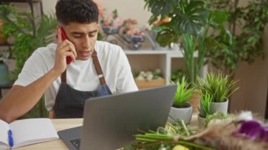 Çiçekçide çalışan, telefonla konuşan ve çeşitli bitkiler arasında dizüstü bilgisayar kullanan genç bir adam..