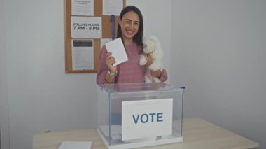Gülümseyen genç bir kadın kapalı bir seçim kolejinde bichon köpeğini tutarken oy veriyor..
