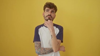 Düşünceli genç İspanyol bir adam, eli çenesinde ve düşünceli bir ifade, izole edilmiş sarı bir arka plana karşı bir soru üzerinde duruyor ve düşünüyor