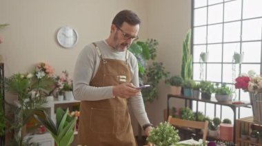 Çiçekçide akıllı telefon kullanan önlüklü, olgun sakallı bir adam.