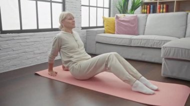 Sarışın bir kadın, oturma odasında yoga yapıyor. Rahat bir kanepesi ve parke zemini var..