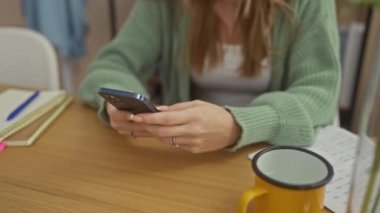 Kapalı bir evde elinde kupa, defter ve çiçekle telefonuyla mesajlaşan genç bir kadın.