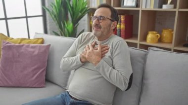Minnettar orta yaşlı bir adam, süveter giyiyor, evde içten bir gülümseme içinde. Göğsündeki eller, sağlıklı bir jest ve sağlıklı bir yüzle parlıyor.