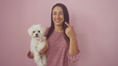 İzole edilmiş pembe arka planda mutlu köpeğiyle dikilirken parmaklarıyla işaret eden genç, gülümseyen İspanyol kadın.