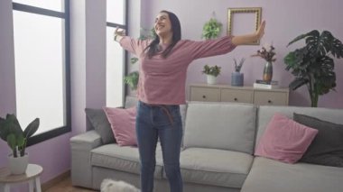 Neşeli bir kadın, sıcacık bir oturma odasında, bichon köpeğiyle kollarını uzatıyor.