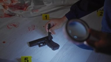 Afrikalı Amerikalı müfettiş olay yerinde üzerinde kanıt numaraları, kan lekeleri ve ön planda büyüteç bulunan bir tabancayı inceliyor..