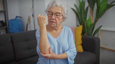 Yaşlı kadın kolunu modern bir oturma odasında muayene ediyor.