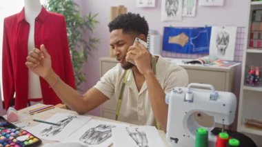 Tasarım çizimleri, kumaş ve dikiş makinesiyle çevrili canlı bir terzi dükkanında telefonla konuşan bir adam..
