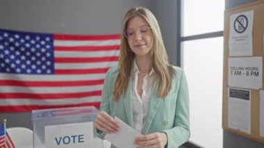 Genç sarışın bir kadın, Amerikan bayrağıyla sarılı bir seçim odasında oy verirken gülümsüyor..