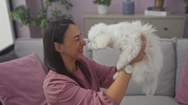 İspanyol kadın elinde beyaz bichon köpeğiyle mor kanepede gülüyor.