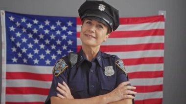 Kendinden emin, orta yaşlı, polis üniformalı bir kadın Amerikan bayrağı önünde kollarını kavuşturuyor..