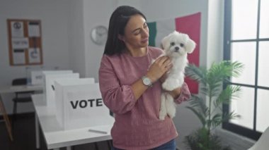 Hispanic woman holding a bichon maltes dog in an indoor college polling station with a banner.