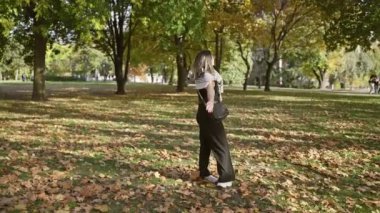 Gözlüklü ve omuz çantalı neşeli genç bir kadın huzurlu bir parkta sonbahar yapraklarının arasında duruyor..