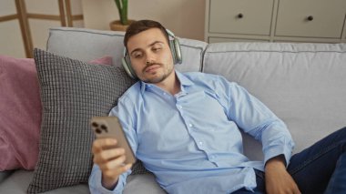 Sakallı, kulaklık takan, mavi gömlekli, oturma odasındaki bir kanepede dinlenen ev ortamında telefonuna bakan yakışıklı İspanyol bir adam..