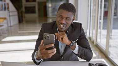 Modern ofis ortamında akıllı telefon kullanırken gülümseyen takım elbiseli Afrikalı Amerikalı bir adam.