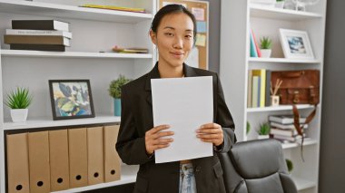 Profesyonel bir Asyalı kadın modern ofis ortamında iş kıyafetlerini gösteren bir belge tutuyor..