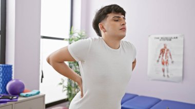 Fizik tedavi kliniğinde egzersiz aletleri ve anatomi posterleriyle sırt ağrısı çeken bir adam..