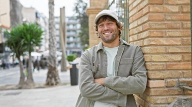 Gülümseyen, sakallı, genç beyaz bir adam. Açık havada, canlı bir şehir caddesinde kollarını kavuşturdu.