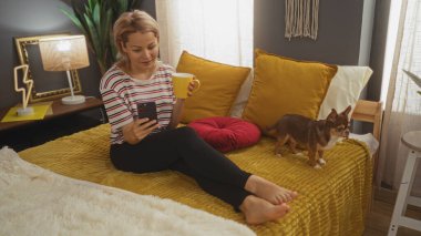 Genç sarışın bir kadın, rahat bir yatak odasında bir chihuahua köpeğiyle, akıllı telefon kullanıyor ve canlı yastıklarla dolu bir yatakta otururken elinde sarı bir bardak tutuyor..