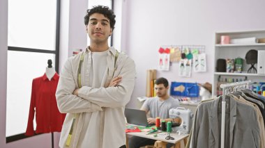 İki adam, kendine güvenen bir terzi ve meslektaşı iyi donanımlı atölyelerinde çalışıyorlar, etrafları giysi ve dikiş ekipmanlarıyla çevrili..