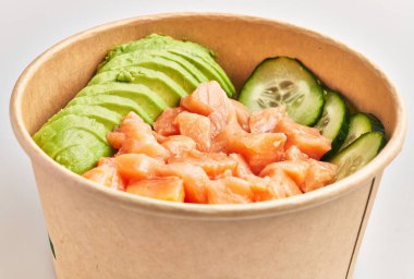 Dilimlenmiş avokado ve salatalıklı somon beyaz arka planda bir kağıt kasede..