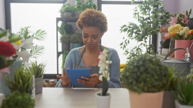 Bitkilerle ve doğal ışıkla dolu bir çiçekçide tablet kullanan Afrikalı bir kadının portresi.