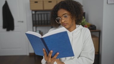 Güzel, kıvırcık saçlı, genç, Afro-Amerikalı bir kadın ofis ortamında mavi kitap okuyor, çekici ve düşünceli bir atmosfer yaratıyor..