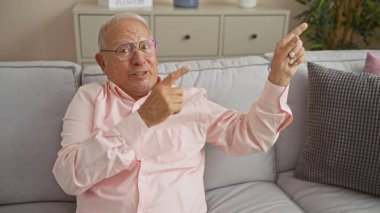 Beyaz saçlı, pembe tişörtlü yaşlı beyaz bir adam oturma odasındaki gri bir kanepede otururken neşeli bir şekilde işaret ediyor..