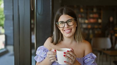 Neşeli, güzel İspanyol kadın bir kafe masasında sabah kahvesinin keyfini çıkarıyor. Sıcak içkisini rahat bir gülümsemeyle yudumlarken, gözlükleriyle ışıltılı pozitifliğini vurguluyor.