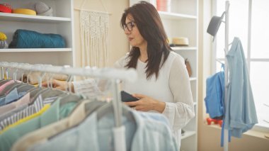 Olgun İspanyol kadın, elinde akıllı bir telefonla kapalı bir gardırop odasında kıyafetlerini karıştırıyor..