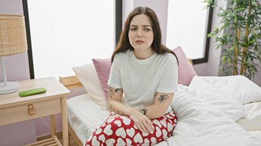 Üzerinde dövmeleri olan düşünceli genç bir kadın rahat bir yatak odasındaki kalp desenli pijamalarıyla bir yatakta oturuyor..