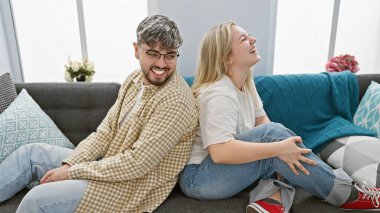 Modern bir oturma odasında sırt sırta oturan gülen çift, gündelik sevgi ve rahatlığı somutlaştırıyor..