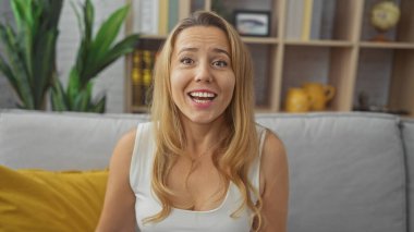 Sarışın kadın, rahat bir oturma odasında modern dekorla rahatça gülümsüyor, rahatlatıcı ve davetkar bir hava yayıyor..