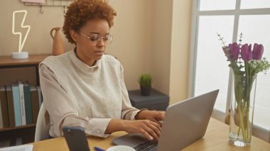 Odaklanmış bir Afrikalı Amerikalı kadın iyi aydınlatılmış bir ofis ortamında dizüstü bilgisayarla çalışıyor..