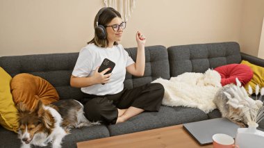 Kanepede köpeğiyle dans eden İspanyol bir kadın, dizüstü bilgisayar, evde tek başına müzik dinliyor..